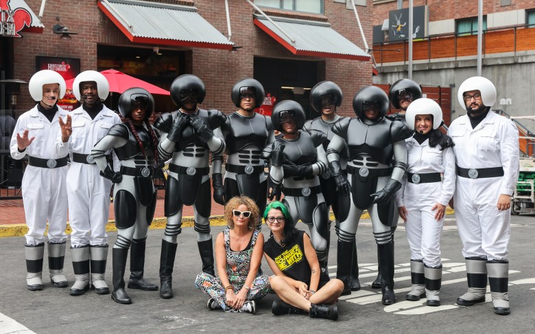 Two people sit with an array of people in black and white armor with round helmets stand behind them