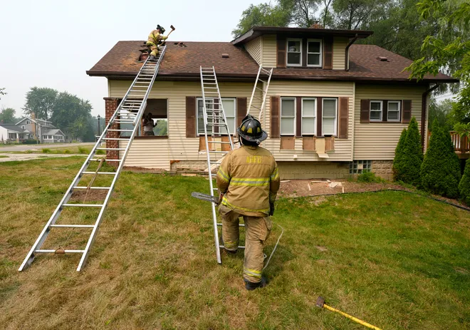 From left, Auburn Hills firefighter Logan Young works at poking a hole in the roof of a house as firefighter Edwin Prado looks on during a training exercise on Auburn Road in Auburn Hills on Thursday, July 16, 2026.