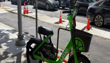 A green LimeGlider electric bike with a basket is locked to a bike rack on a city sidewalk next to a street with cars and an articulated public bus.