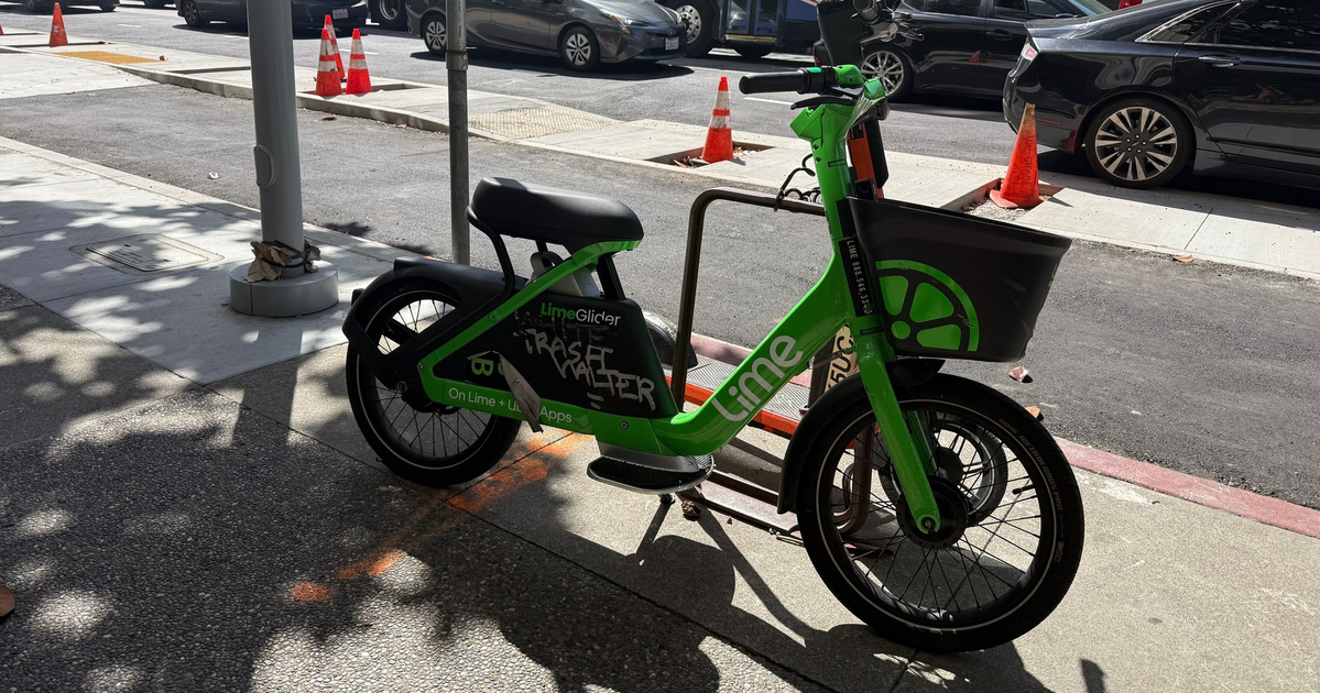 A green LimeGlider electric bike with a basket is locked to a bike rack on a city sidewalk next to a street with cars and an articulated public bus.