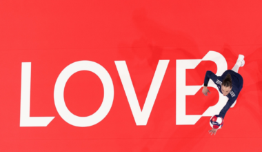 A volleyball player in navy sportswear leaps to hit a ball over a red court with the word "LOVE?" painted in large white letters.