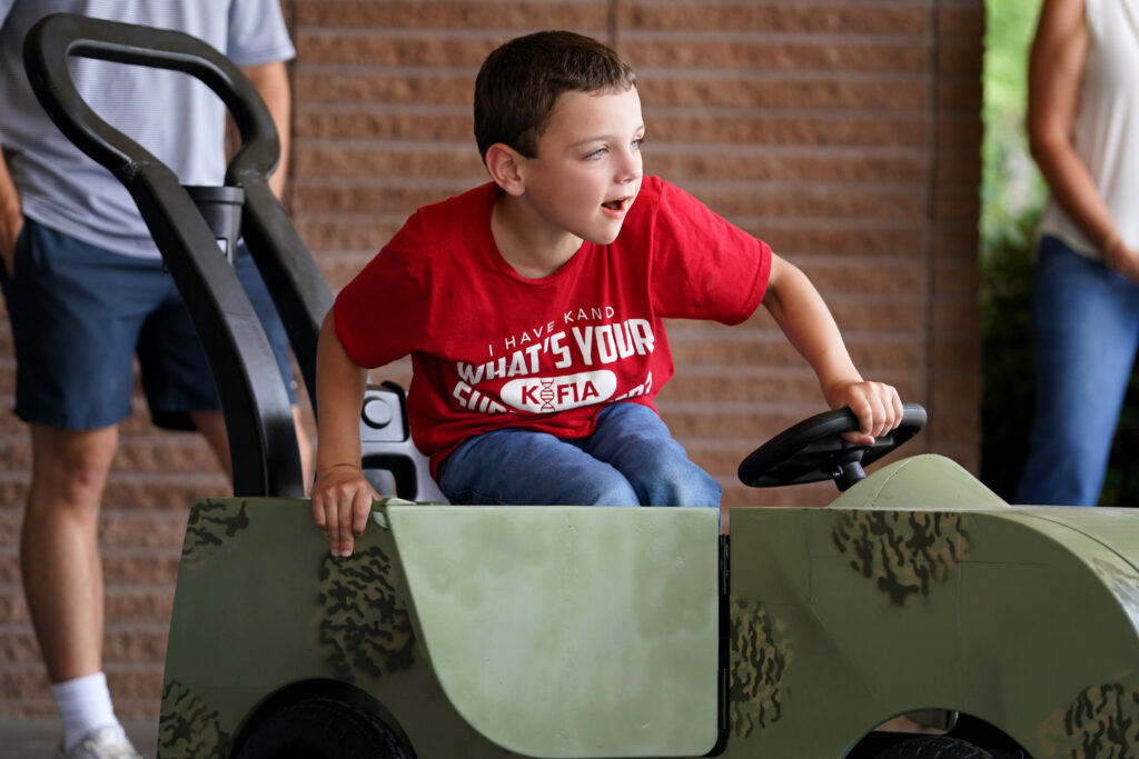 Beau Bewarder sits in his custom-built camouflage ride-on toy car, leaning forward with excitement as he grips the steering wheel.