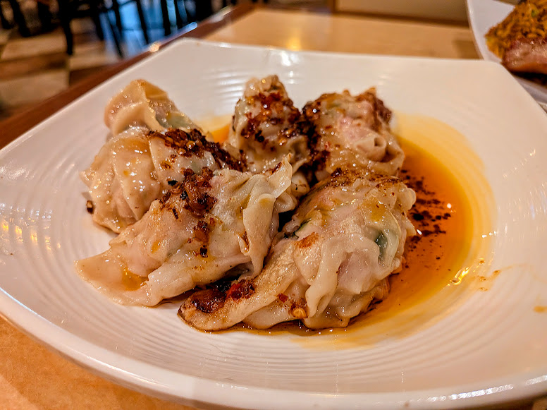 Four steamed dumplings topped with chili oil and seasonings are served on a white plate.