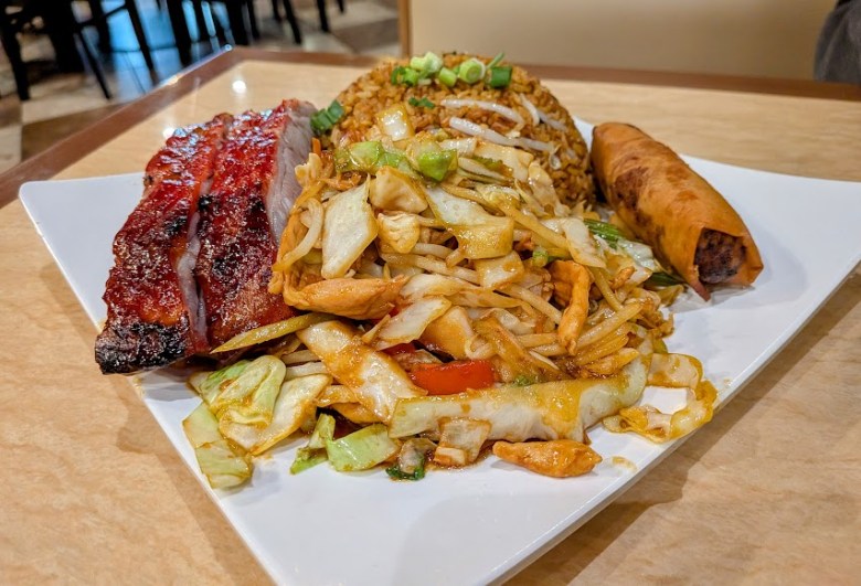 A plate with barbecue pork ribs, stir-fried vegetables with chicken, fried rice topped with green onions, and a spring roll on a white square dish.