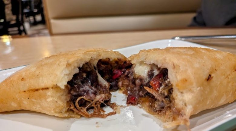 A fried empanada cut in half on a white plate, showing a filling of shredded meat, black beans, red peppers, and melted cheese.