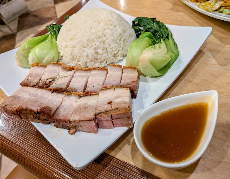 A plate of sliced roast pork belly, steamed white rice, bok choy, and a small dish of brown dipping sauce on the side.