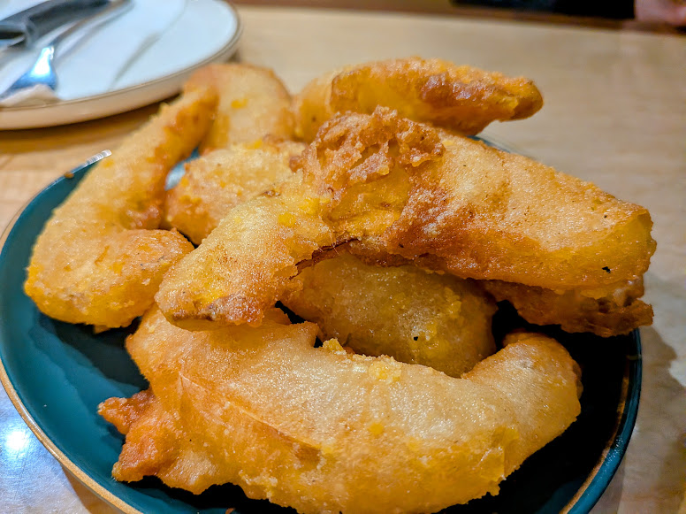 A plate of golden-brown battered and fried fish fillets sits on a table next to a plate with cutlery.
