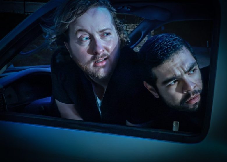 Two men sit in a car at night, leaning out of the window and looking intently in the same direction with serious expressions.