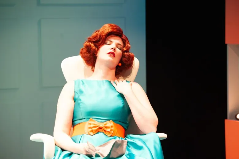 A person with red hair in a retro teal dress and orange belt sits in a chair with eyes closed, head tilted back, and hand on their chest.