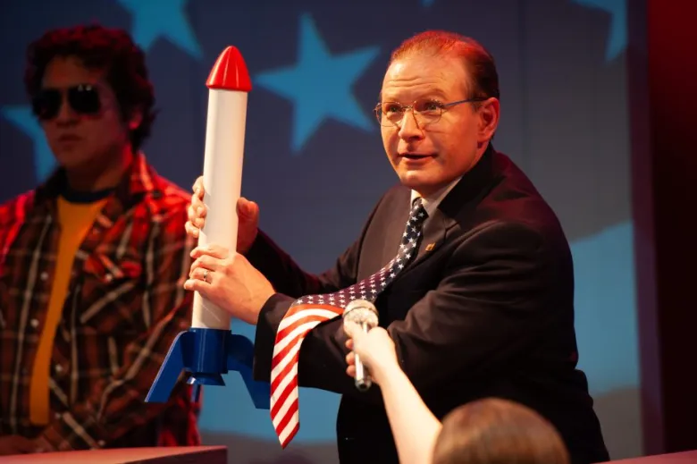 A man in a suit and American flag tie holds a toy rocket while someone holds a microphone toward him; another person stands in the background wearing sunglasses.