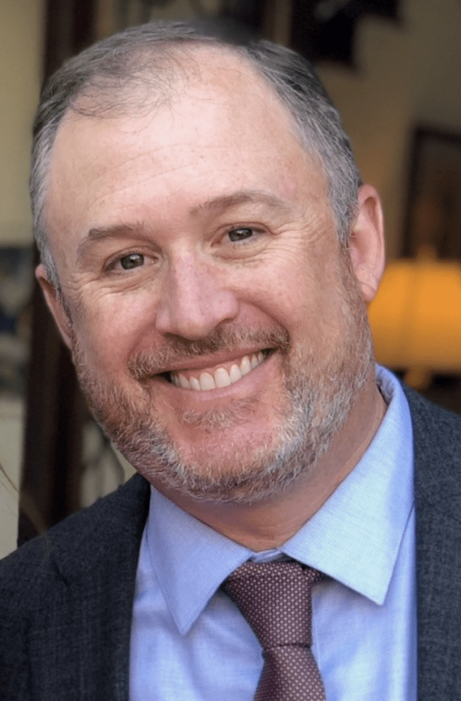 A white man with grey hair on his temples and his beard. He is wearing a dark suit with a blue shirt and colorful tie.
