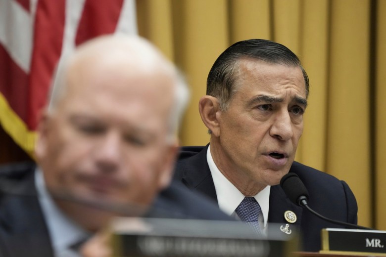 Rep. Darrell Issa speaks into a microphone at an official hearing, with another man blurred in the foreground.