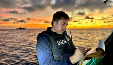 Drew LoVullo, in a U.S. Coast vest, writes on a paper while aboard a ship with the sunset in the background.