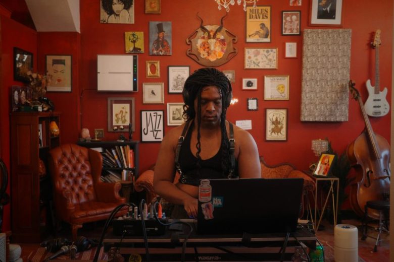 A person with braided hair sits at a laptop in a red room decorated with framed art, musical instruments, and bookshelves.