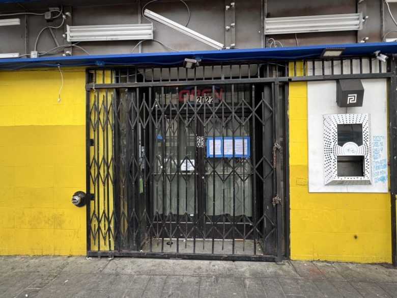 A black metal gate covers a glass door with papers posted on it, set in a yellow wall beside an ATM.