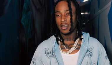 A young Black man in a light blue button-up shirt over a white tank top stands between two walls in the nighttime, looking downward and to the left