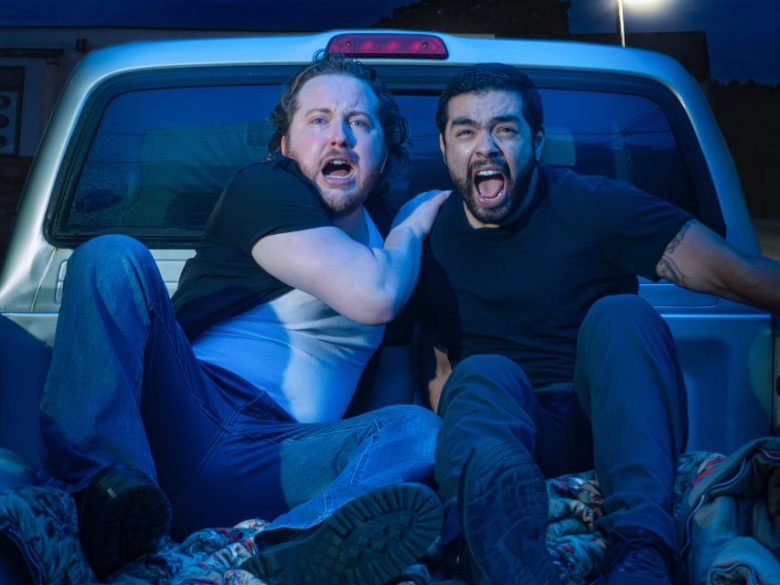 Two men sit in the back of a pickup truck at night, both with wide eyes and open mouths, appearing startled or scared.