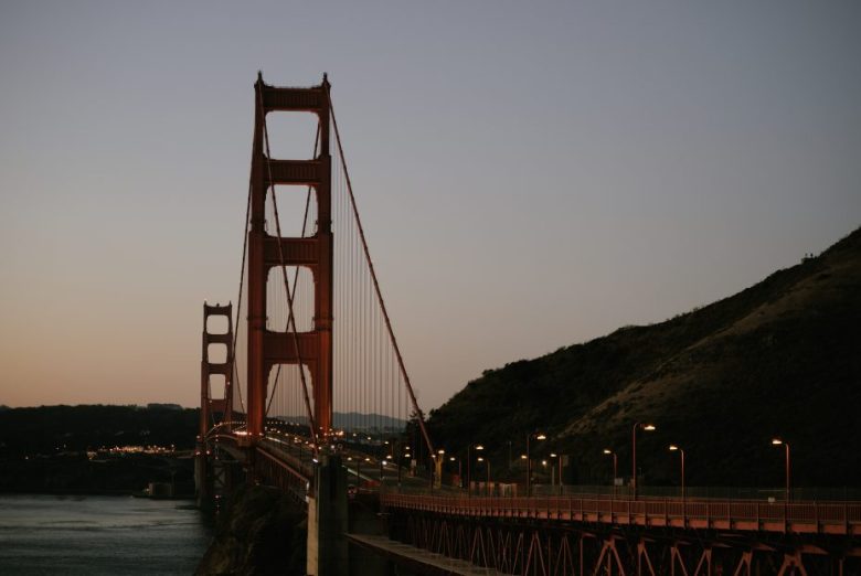 Golden Gate Bridge at dusk with lit streetlamps, spanning a body of water and leading to a hill.