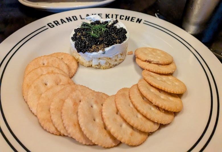 Round crackers are arranged in a curve on a plate around a small stack topped with black caviar and a sprig of dill. The plate says "GRAND LAKE KITCHEN.