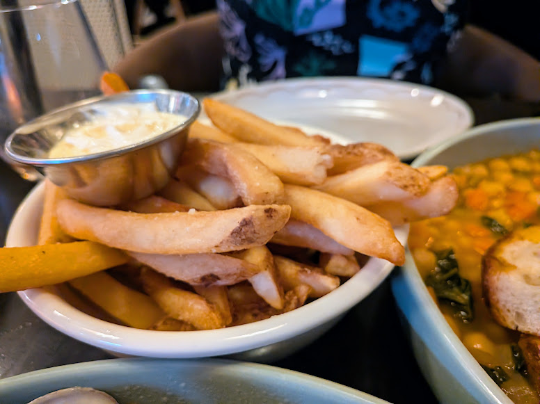 A bowl of thick-cut French fries with a metal cup of dipping sauce, next to a bowl of stew and bread, on a restaurant table.