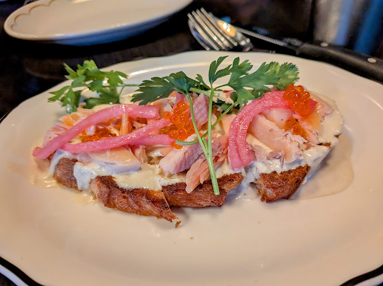Open-faced sandwich with shredded chicken, creamy sauce, pickled onions, fish roe, and parsley on a slice of bread, served on a white plate with a fork and knife in the background.