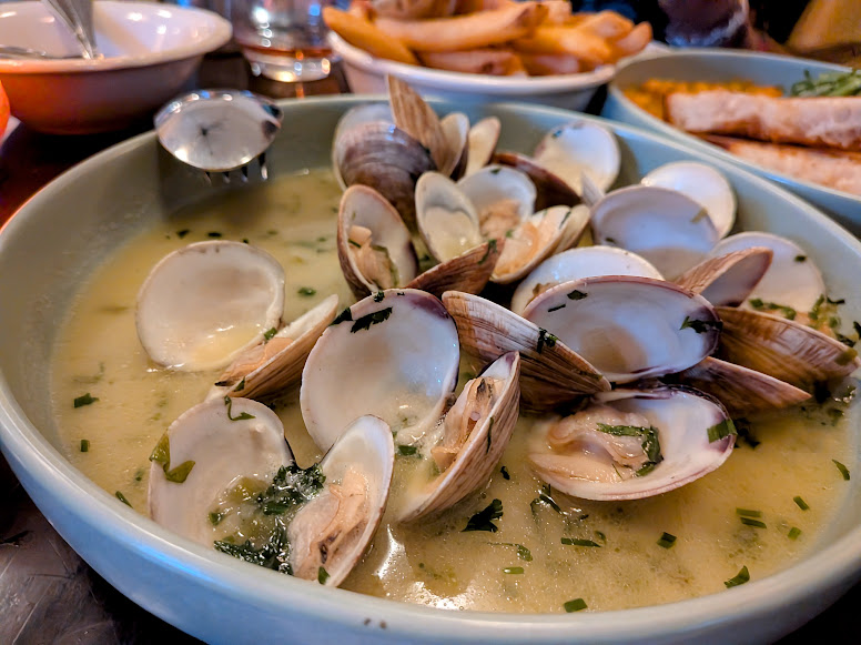 A bowl of clam chowder with open clam shells in broth, garnished with herbs. Fries and bread are visible in the background.