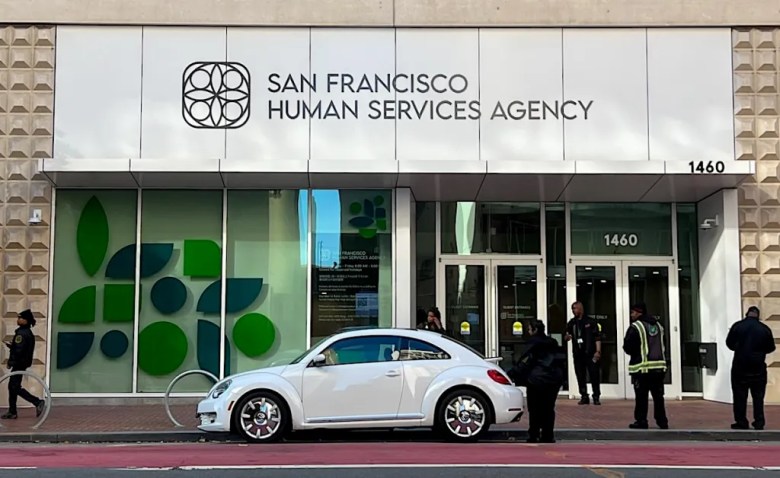 a white VW Bug in front of a building with a sign reading "San Francisco Human Services Agency" with several security guards nearby. 