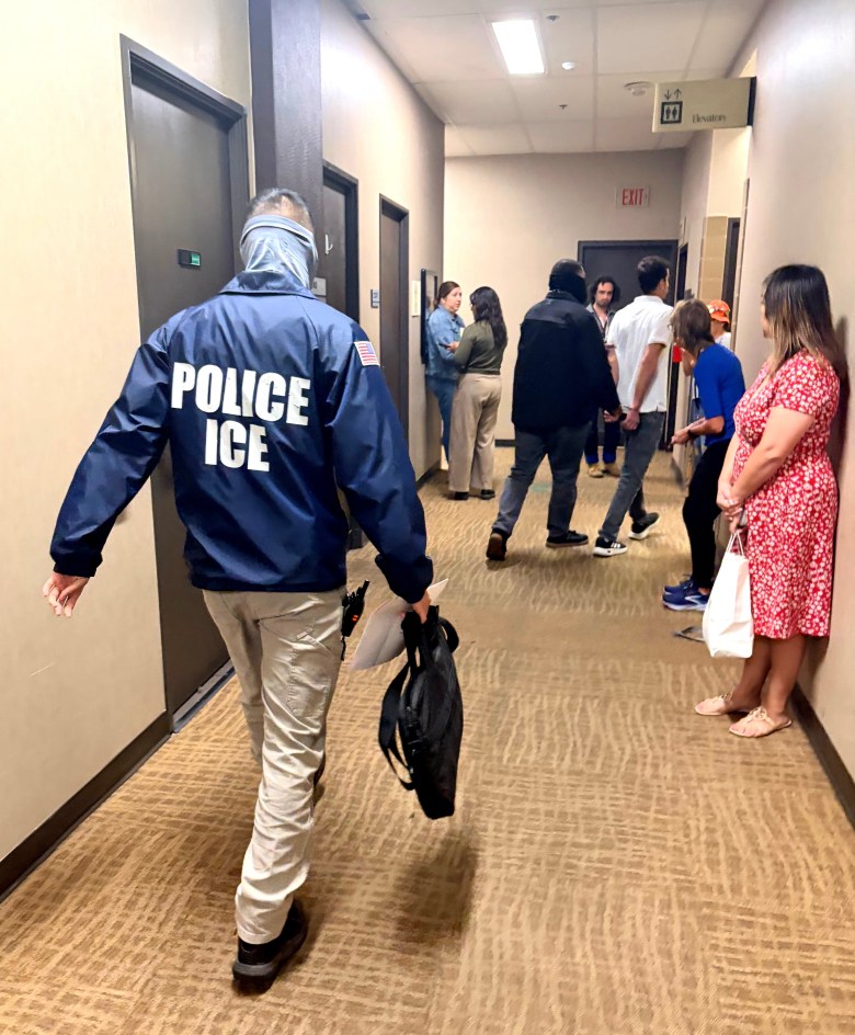 People stand in a hallway ahead of a person wearing a jacket labeled "POLICE ICE"
