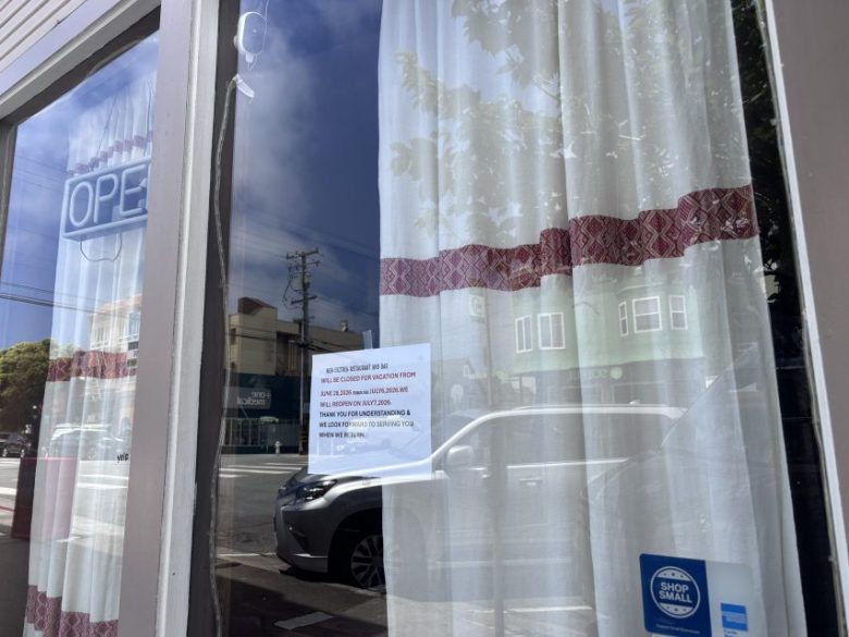 A storefront window with white curtains, an "OPEN" sign, and a printed notice taped to the glass. A car and buildings are reflected in the window.