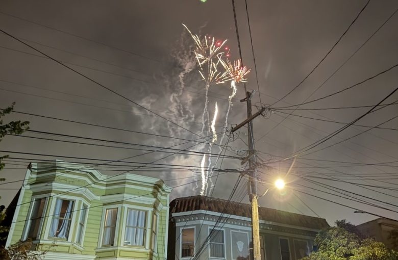 Fireworks explode in the night sky above residential houses, with power lines and a streetlight in the foreground.