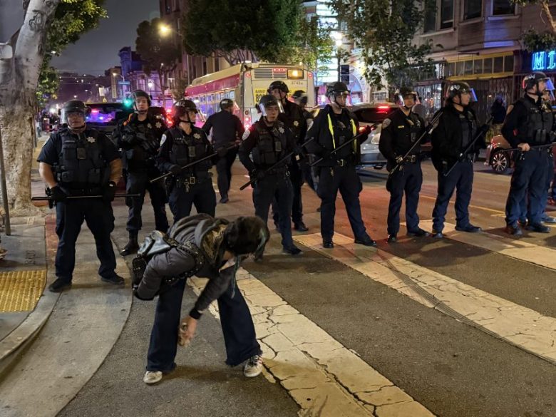 A line of police officers in riot gear stands across a city street at night as a person bends down near a crosswalk in the foreground.