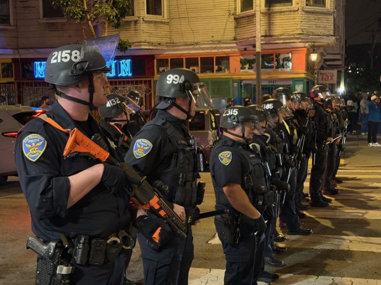 A line of uniformed police officers wearing helmets and holding batons and rifles stands on a city street at night.