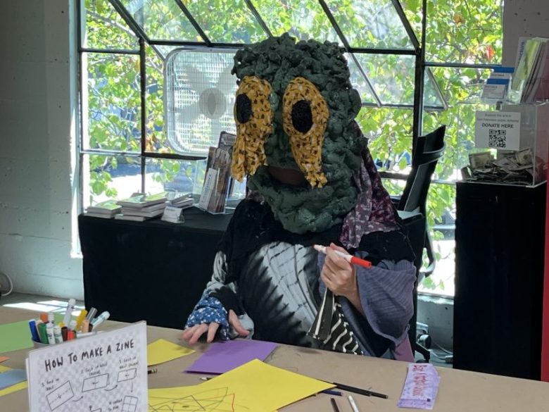 Person wearing a large, green mask with big eyes and a skeleton shirt sits at a table creating art with colored paper and markers. A sign reads "How To Make A Zine.