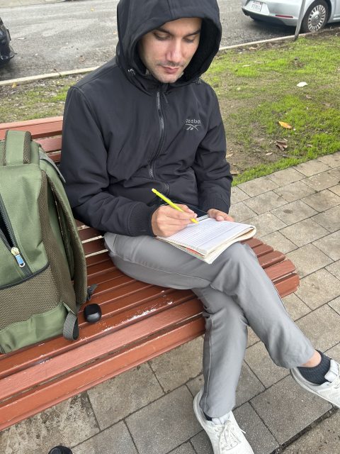 A person in a black hooded jacket sits on a wooden bench, writing in a notebook with a yellow pen. A green backpack is beside them.