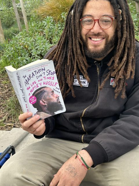 A person with dreadlocks and glasses sits outdoors, smiling and holding the book "The Marathon Don't Stop: The Life and Times of Nipsey Hussle.