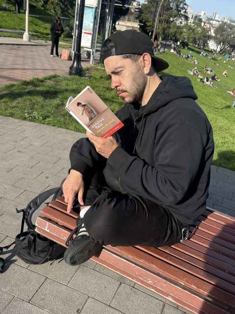 Man in black hoodie and cap sits cross-legged on a bench reading a book outdoors in a park on a sunny day.