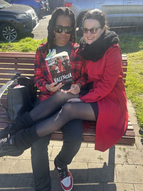 Two people sitting on a bench outdoors; one is holding a book titled "Valencia." Both are wearing sunglasses and red jackets, and sunlight is casting shadows on the ground.