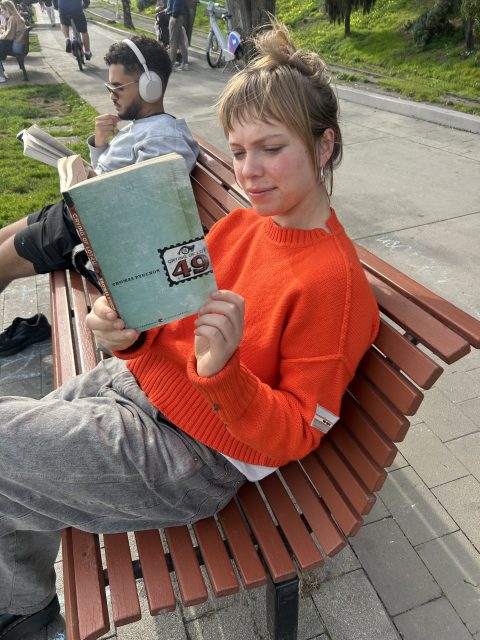 A woman in an orange sweater sits on a bench reading "The Crying of Lot 49" outdoors, with other people relaxing nearby.