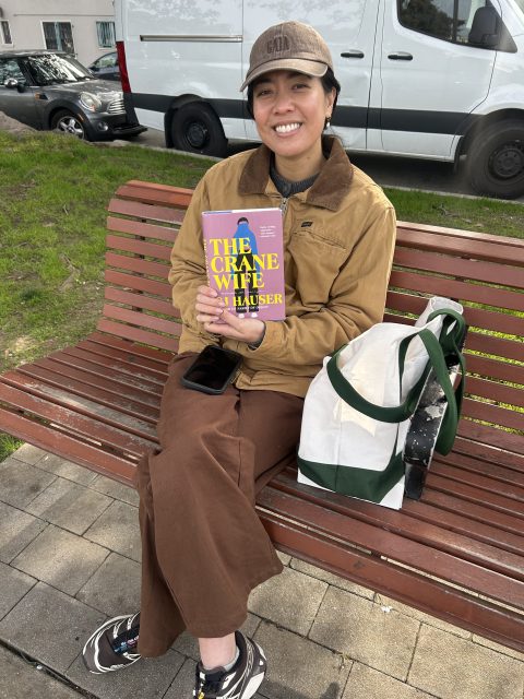 A person sitting on a bench outdoors, smiling and holding a book titled "The Crane Wife" by CJ Hauser, with a tote bag and phone beside them.