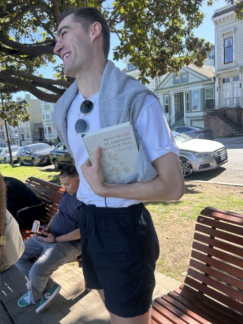 A man stands outdoors near a bench, holding a book and wearing a white t-shirt, black shorts, and a sweatshirt draped over his shoulders. Other people and cars are visible in the background.