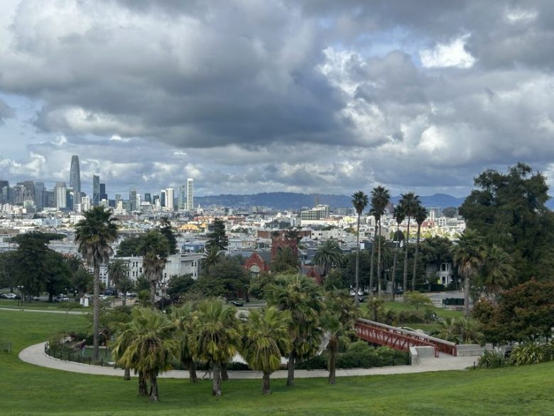 View of a city skyline with tall buildings under cloudy skies, palm trees and a park with a red bridge in the foreground.