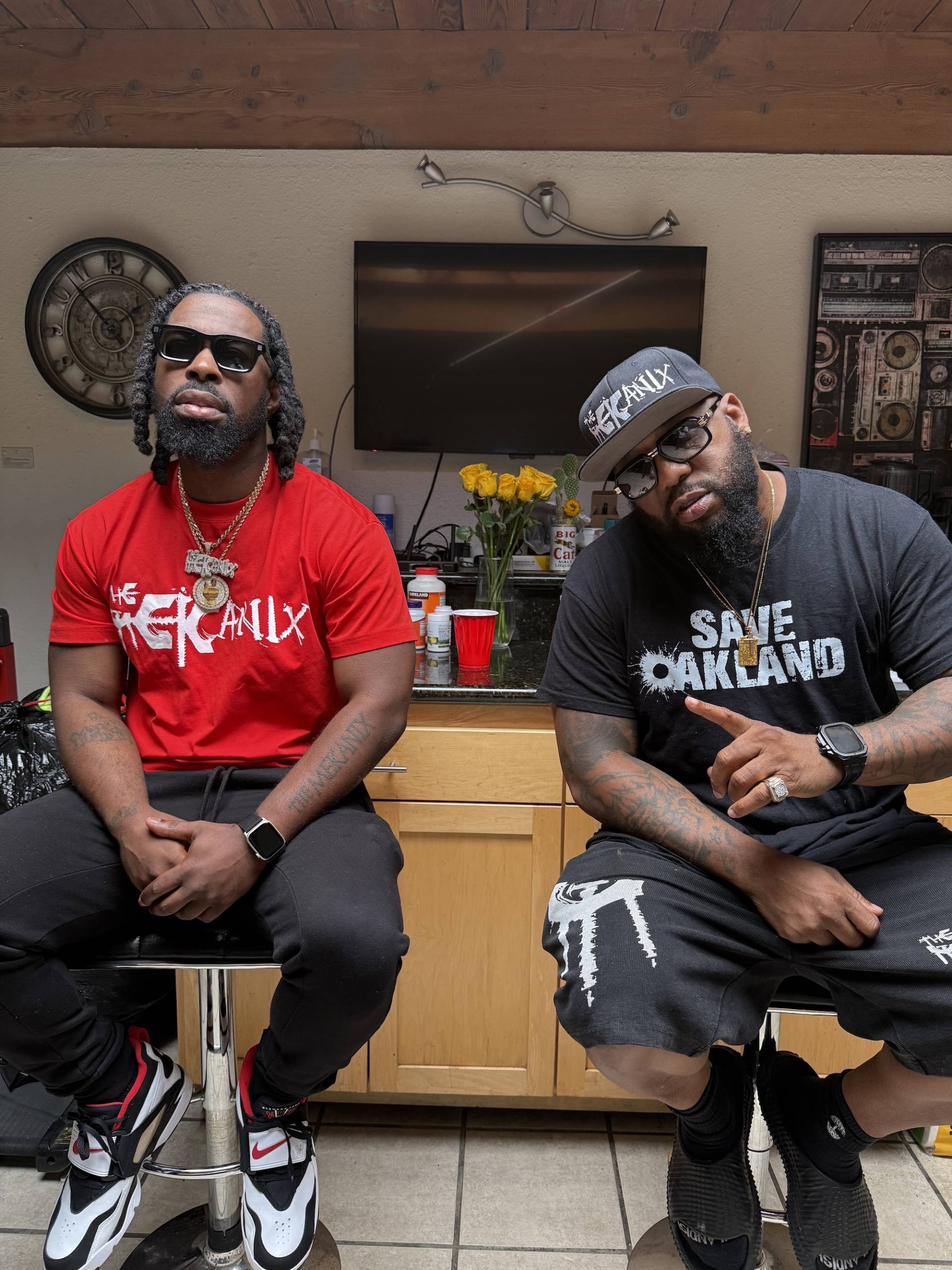 Two men pose for a photo while sitting on bar stools in a music studio.
