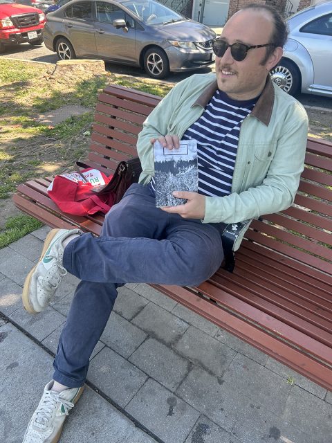 A man wearing sunglasses sits on a wooden bench outdoors, holding an open book. A red bag and a pack of LaCroix are beside him. Cars are parked in the background.
