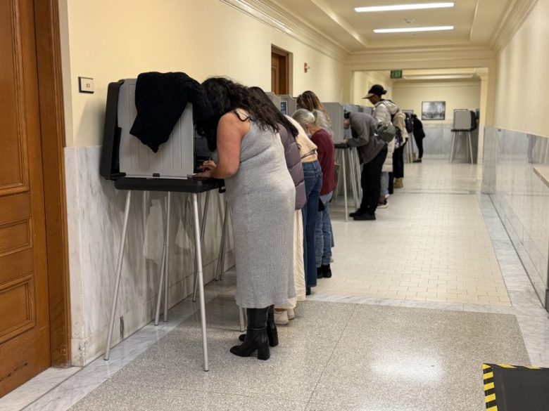People stand in a line at voting booths in a hallway, casting their ballots in an indoor polling place.