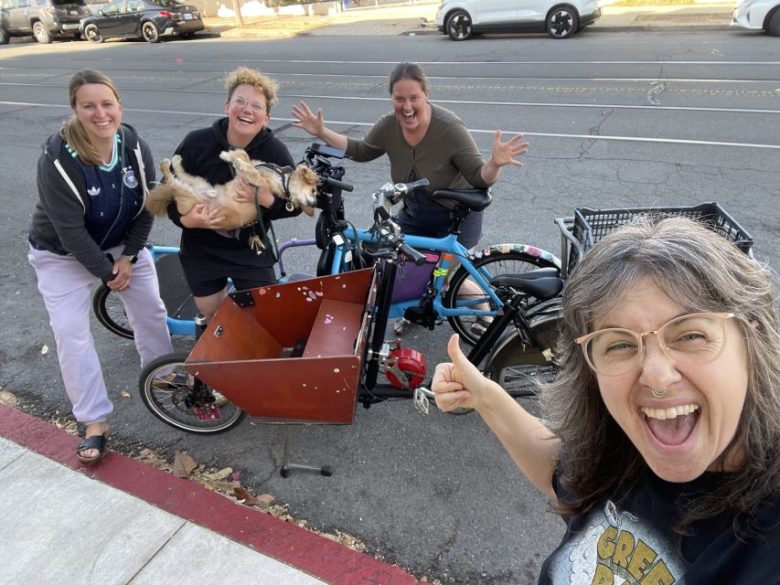 Four smiling women pose with a cargo bike carrying two small dogs along a street; one woman holds up her hand for a selfie.