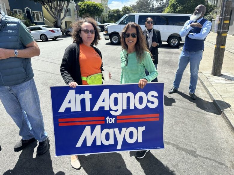 Two women stand on a street holding a blue and white “Art Agnos for Mayor” sign. Three other people stand in the background near parked cars.