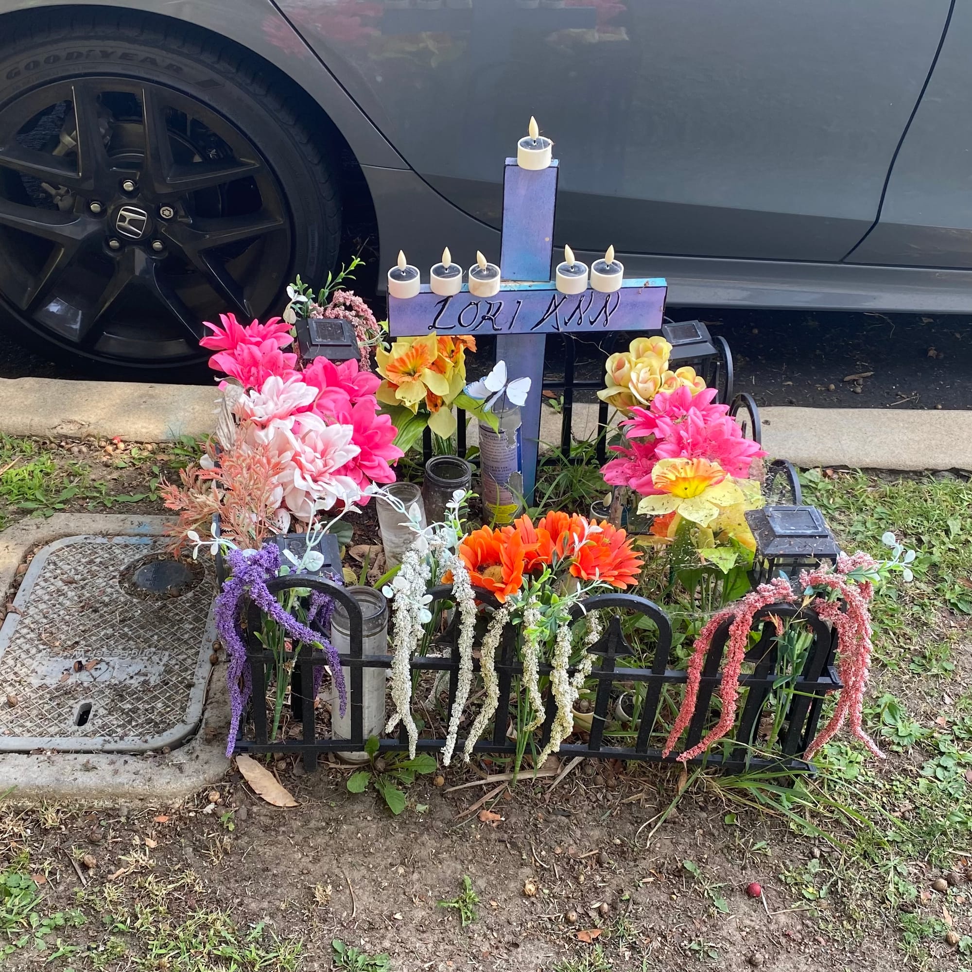 A very low black fence encloses a group of brightly colored flowers, candles and a purple cross bearing the name "Lori Ann" next to a gray car parked on the side of the road.