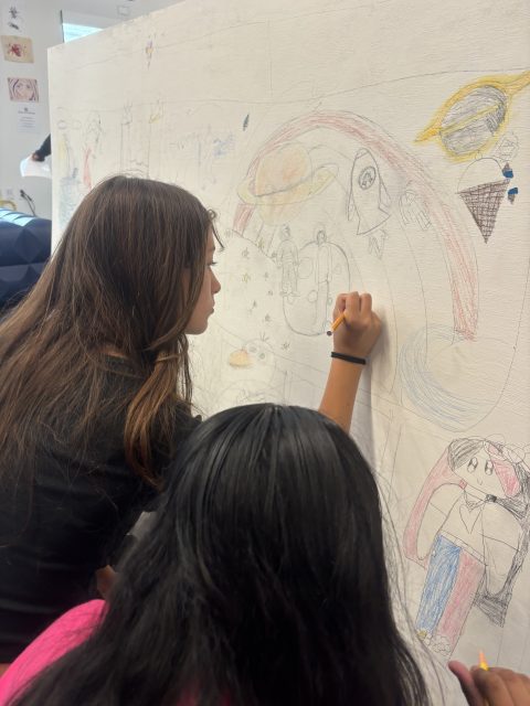 Two girls draw colorful rockets, planets, and rainbows with pencils on a large white wall mural in a classroom setting.