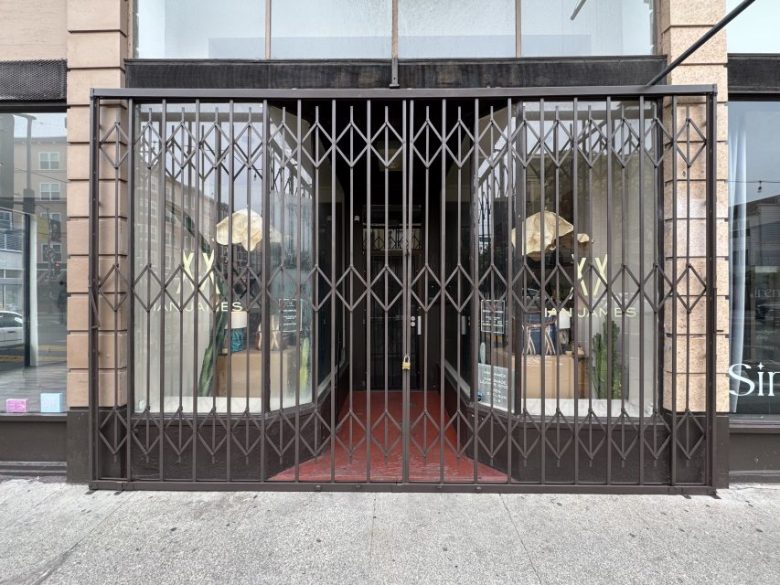 Storefront with metal security gate closed, displaying two mannequins wearing hats in the window displays on either side of a recessed red entryway.