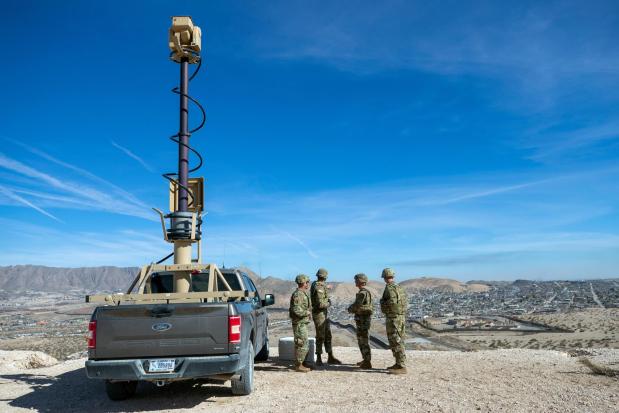 Army soldiers look at the border wall next to a surveillance vehicle.